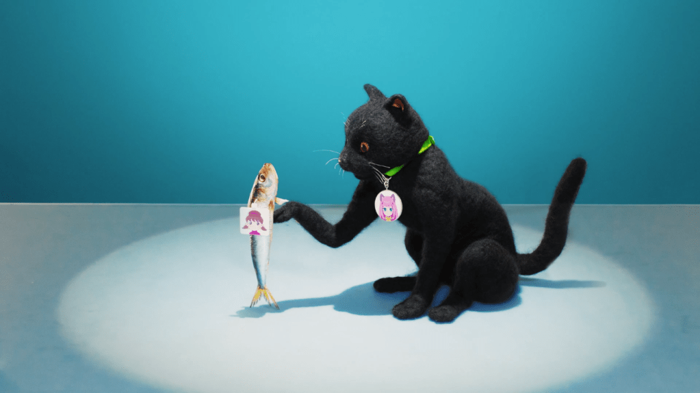 A screencap from Osomatsu-san of a cat puppet (wearing a collar of Nyaa's face) holding a prop fish (tagged with Totoko's face) as if to dance.